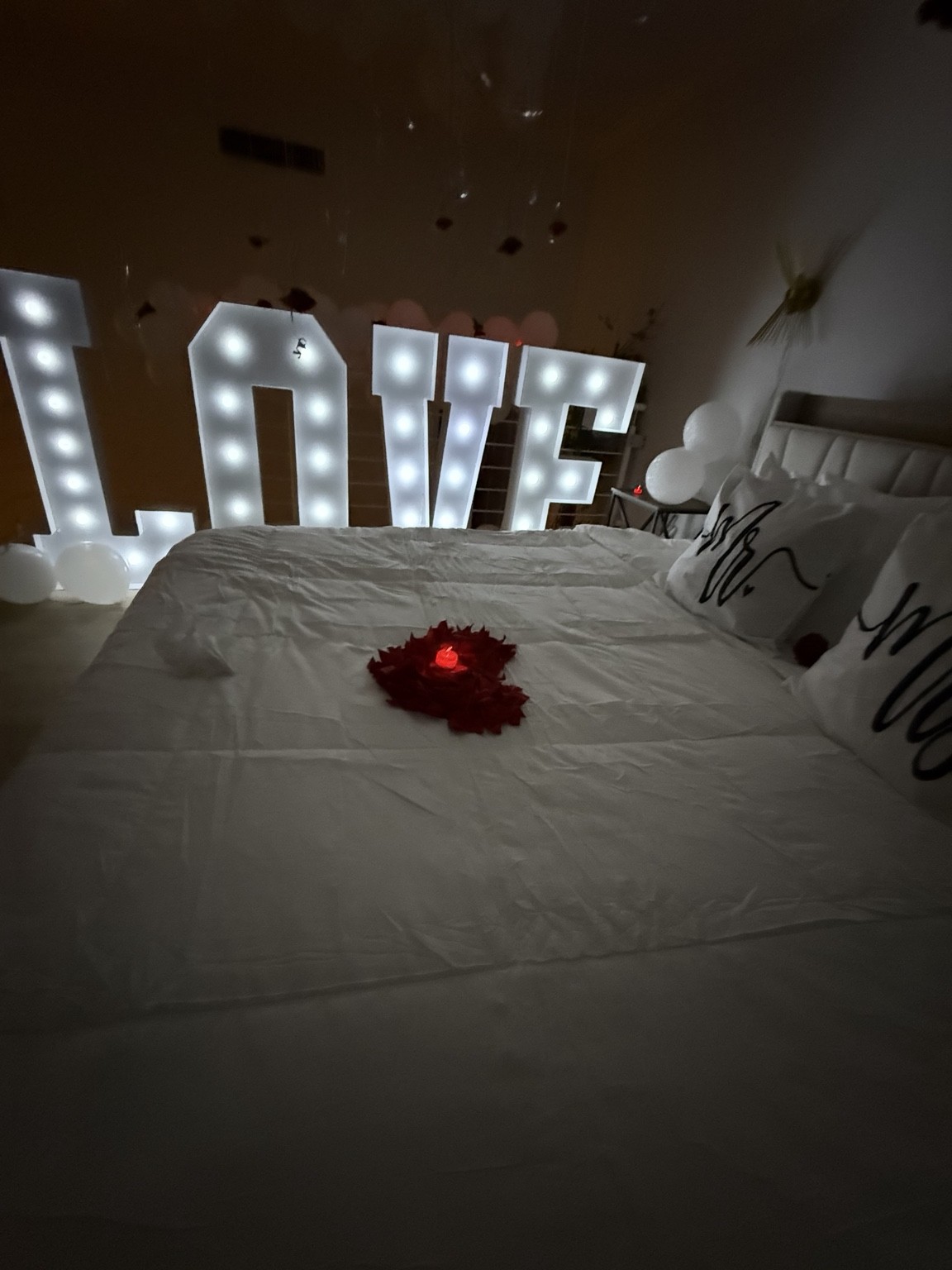 Glowing LOVE letters, rose petals and white bedding create an intimate romantic reveal.